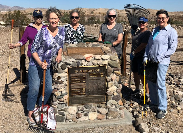 Photograph of six women holding rakes and other tools, gathered around a marker that reads "In Memory Of' with names listed. Taken at the Bullhead City Hardyville Pioneer Cemetery; photo taken by a chapter member and used with permission