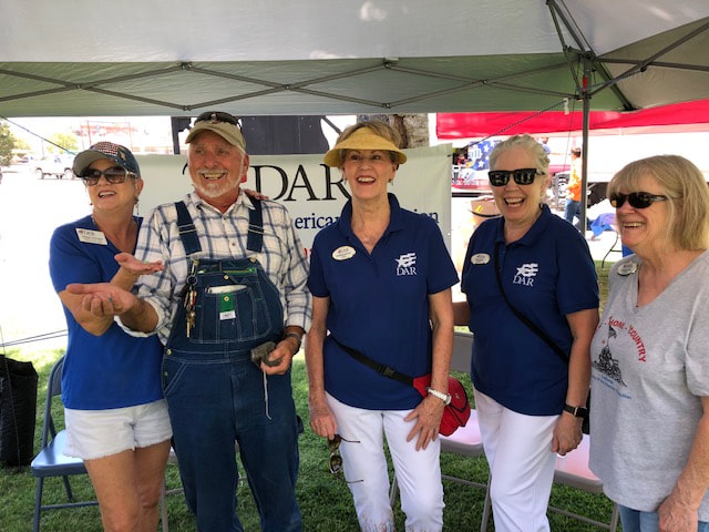 Photograph of a man and four women standing under a white canopy; in the background, a banner reads DAR, Daughters of the American Revolution; photo taken by a chapter member and used with permission